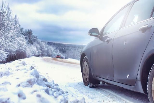A gray car on a snowy road with trees in the background under a cloudy sky.