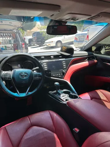Interior of a Toyota Camry. Black steering wheel, red leather seats, center console with infotainment screen. A hat rests on the dashboard.