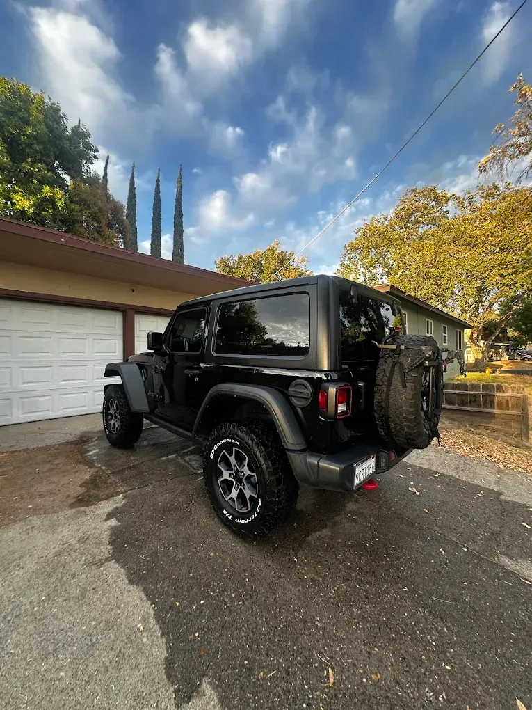 Black Jeep Wrangler parked in front of a garage, under a cloudy blue sky.