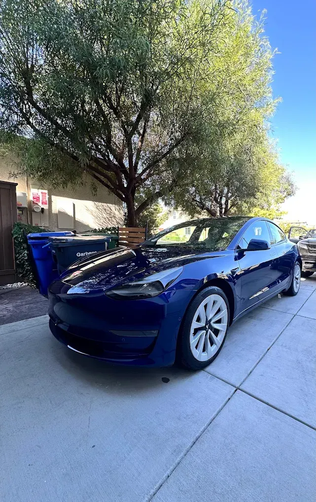 Blue Tesla car parked on a paved driveway with a tree in the background.