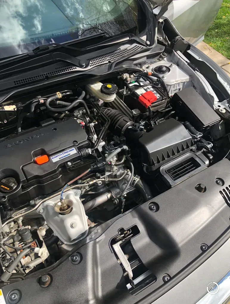 Silver car engine bay, open hood. Black and gray components visible, including battery and air intake.