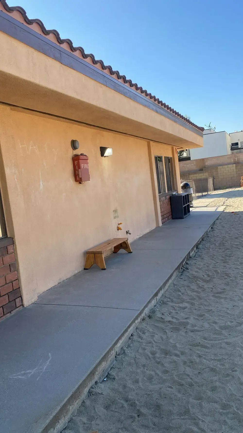 Tan building with a concrete walkway, bench, and red fire extinguisher on the wall. Desert setting.