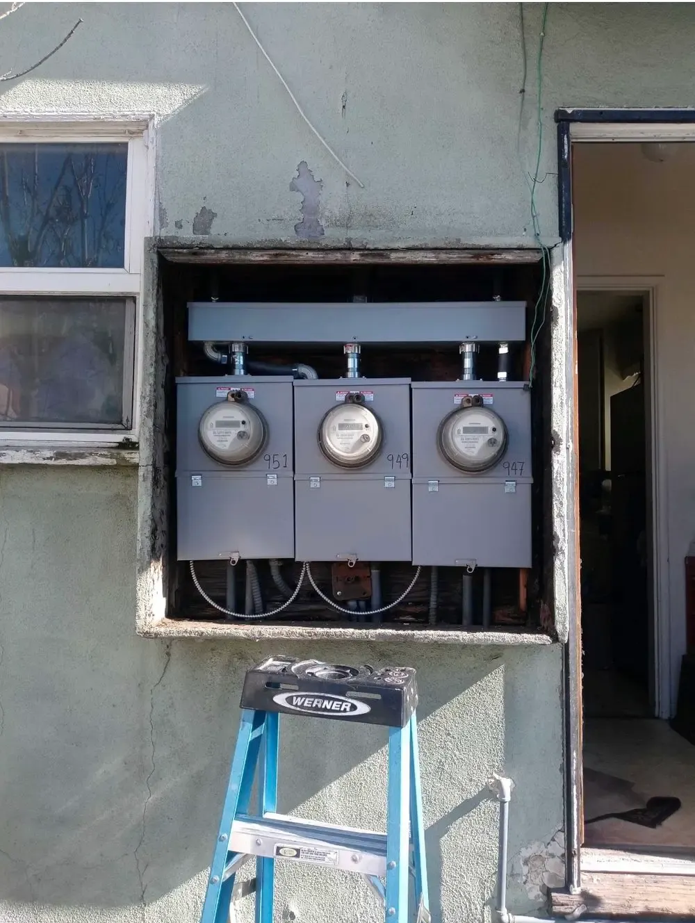 Three electric meters installed in a recessed wall on the exterior of a building. A ladder is in front.