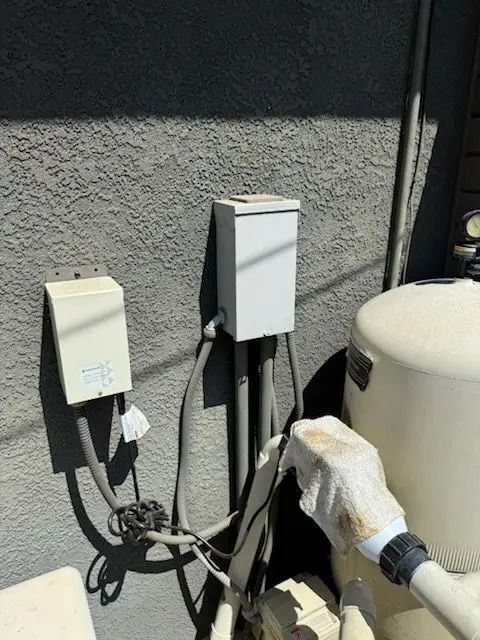 White electrical boxes and conduit on a gray stucco wall near pool equipment.