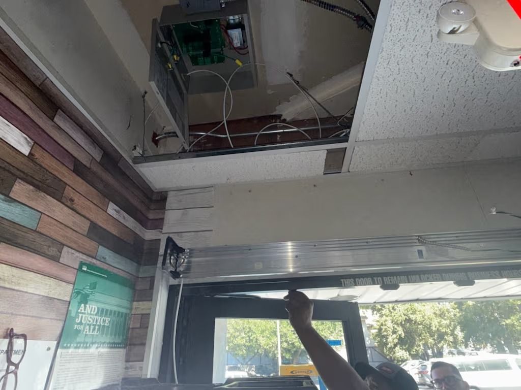 Person working on wiring above a window; exposed ceiling with electrical panel, wood-plank wall.