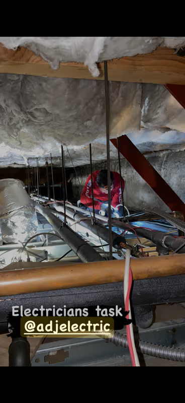 Electrician working in a crawl space with exposed pipes, wiring, and insulation.
