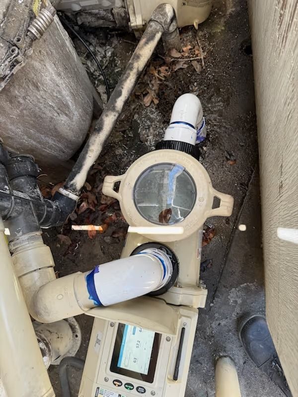 Pool equipment, including a flow meter, piping, and control panel. Beige and black components in a cluttered outdoor setting.