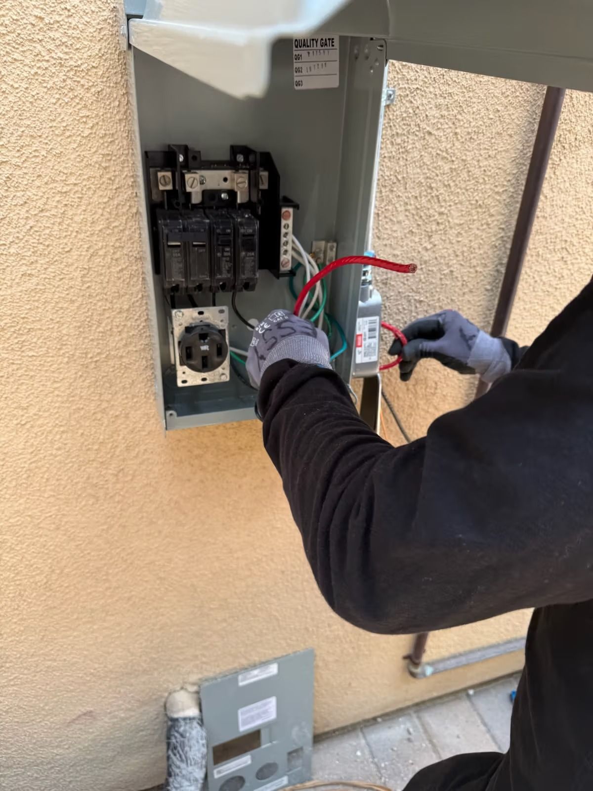 Person in gloves wiring electrical panel on beige stucco wall. Red and green wires are visible.