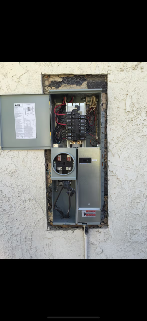 Electrical panel mounted on stucco wall, door open, revealing breakers and meter. Gray metal.