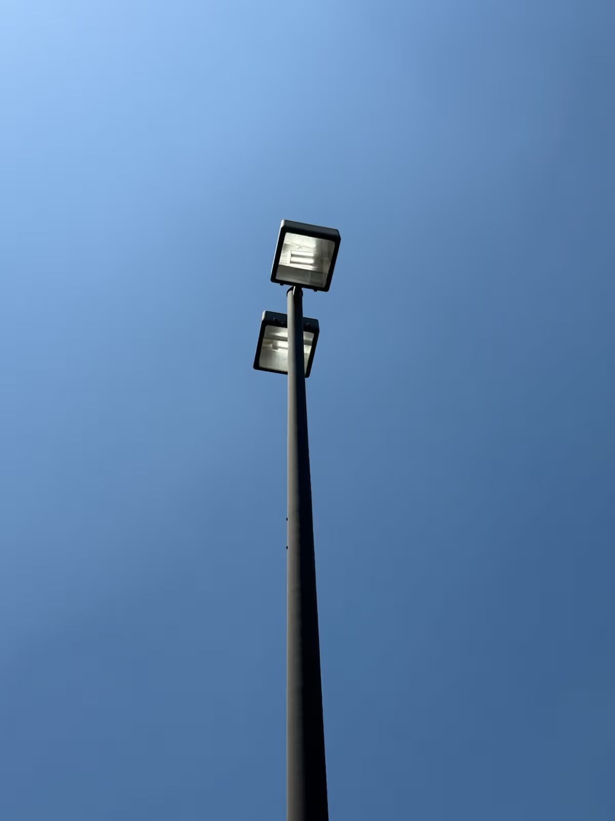 Tall, gray light pole with two rectangular light fixtures against a bright blue sky.