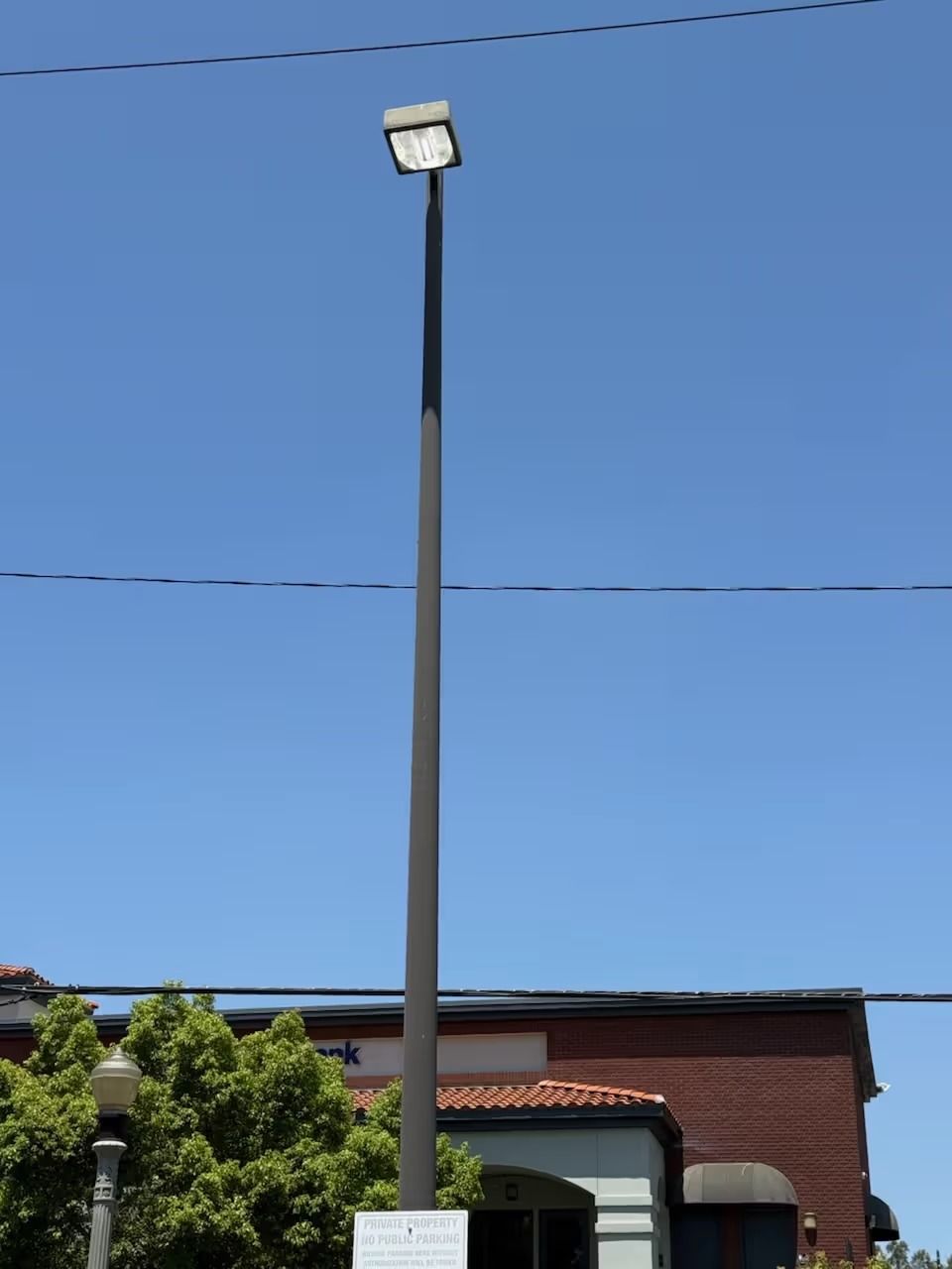 Tall, dark street light with rectangular top against blue sky; trees and building in background.