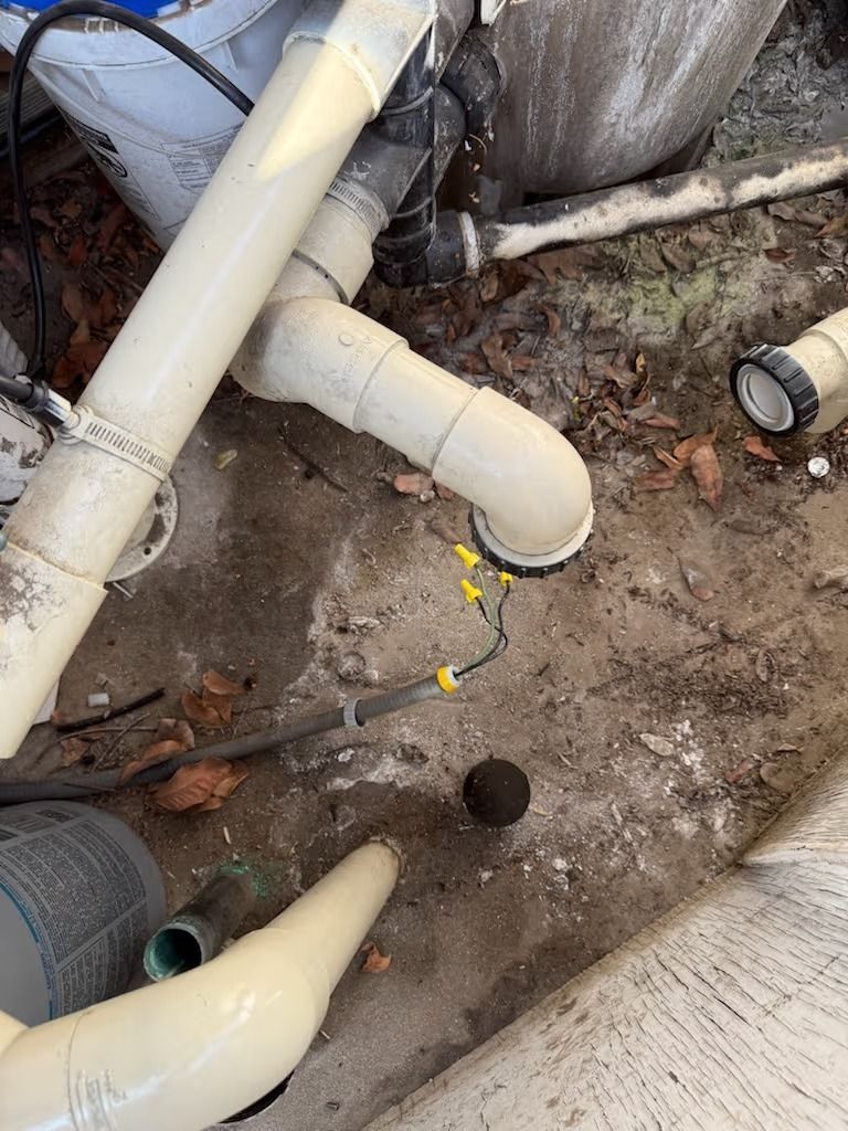 Pool pipes and equipment on concrete ground. Beige pipes, black elbow, wiring, and debris.