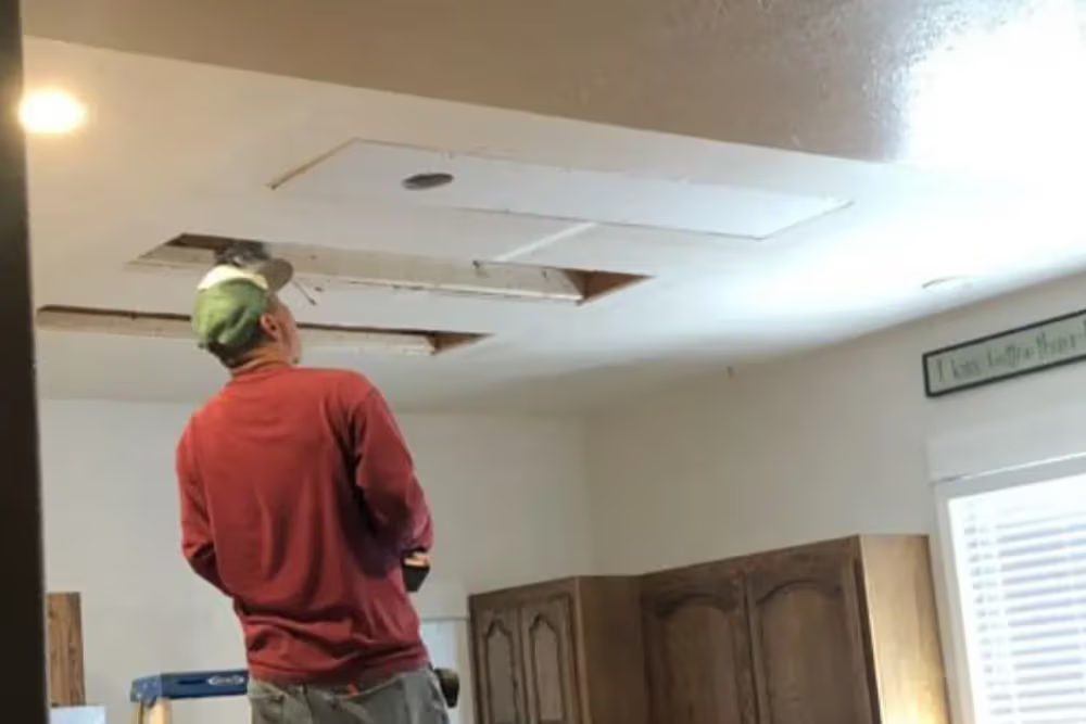 Man on ladder repairs ceiling, removing sections. Kitchen setting, cabinets, natural light.