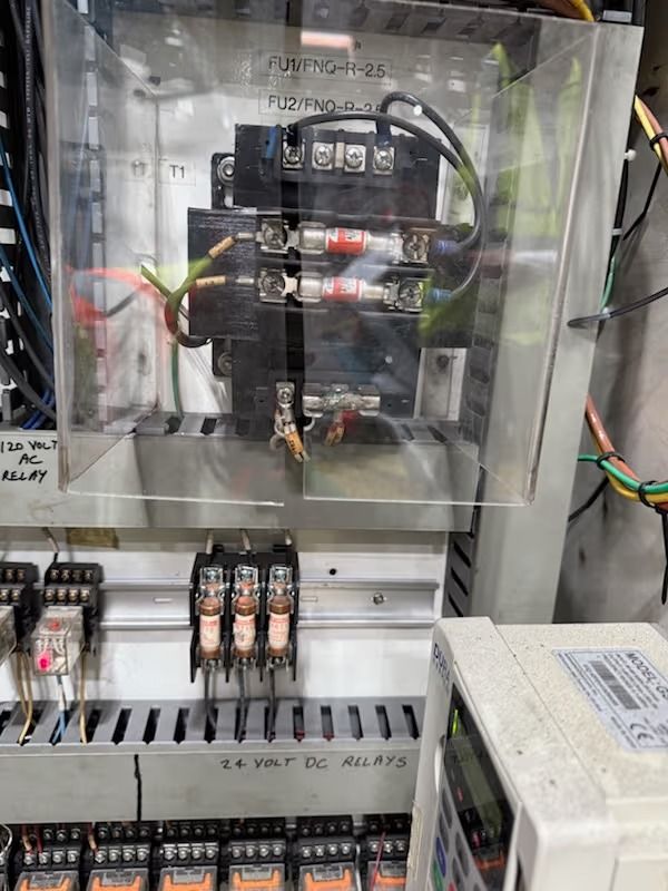 Electrical panel with fuses and wiring, protected by a clear cover, inside a metal cabinet.