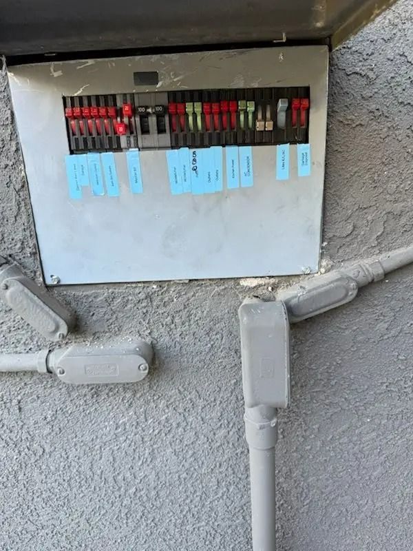 Outdoor electrical panel mounted on a stucco wall. Conduit and connectors are visible. Blue labels identify circuits.