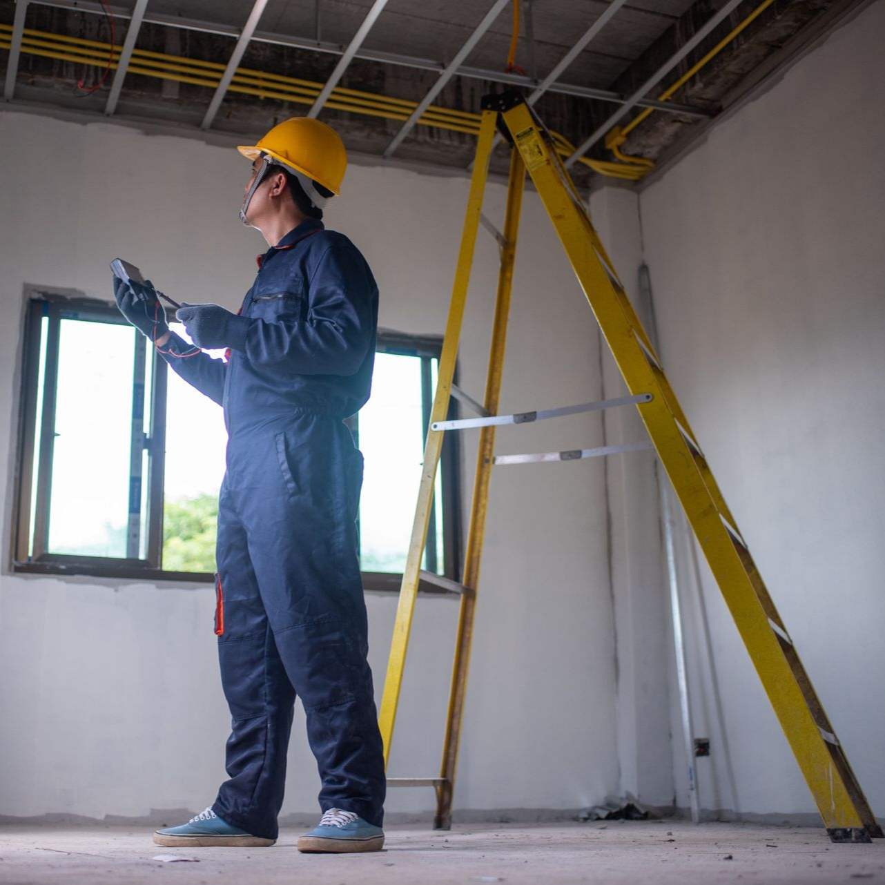 Construction worker in blue jumpsuit and yellow hard hat inspecting wiring near a ladder.