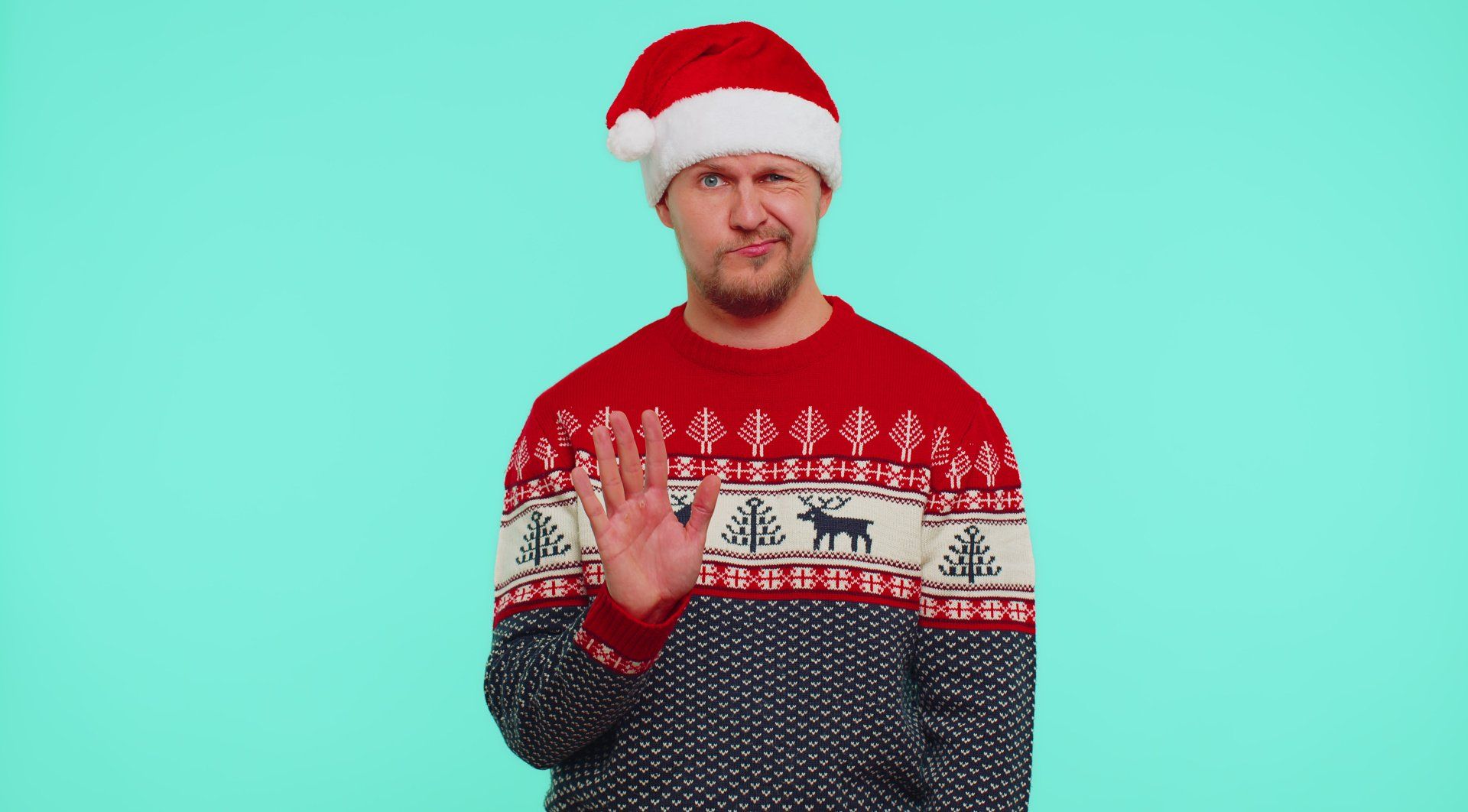 Man in Santa hat and Christmas sweater raising his hand to say no.