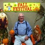 A man is sitting in a chair holding a pumpkin in front of a fall festival sign.