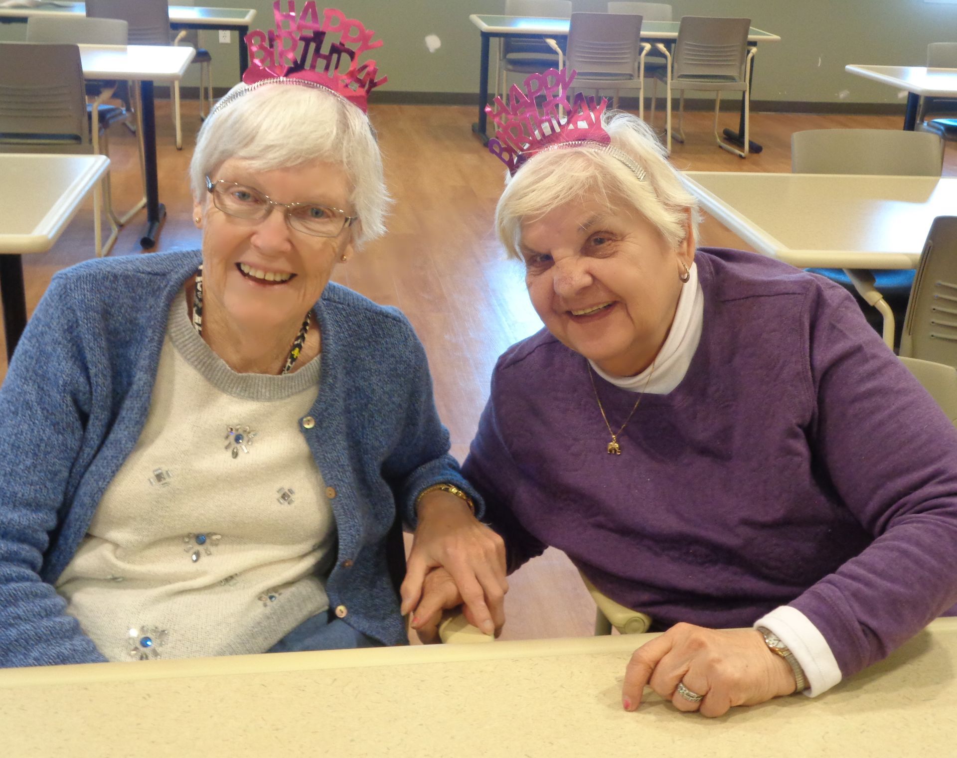 Two elderly women sitting at a table with one wearing a 1st birthday hat