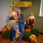 A man is sitting on a scarecrow in front of a fall festival sign.
