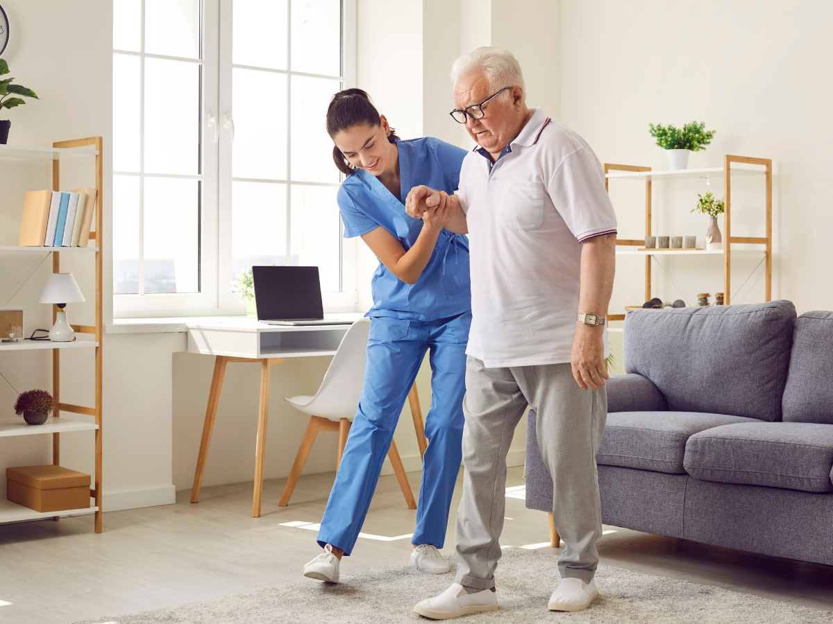 A nurse is helping an elderly man walk in a living room.