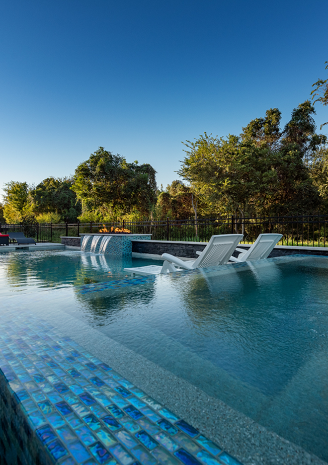 Luxury pool with blue-tiled water feature and white lounge chairs by trees under a clear sky