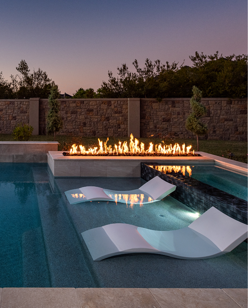Backyard pool at dusk with a glowing fire feature and two white lounge chairs in the water