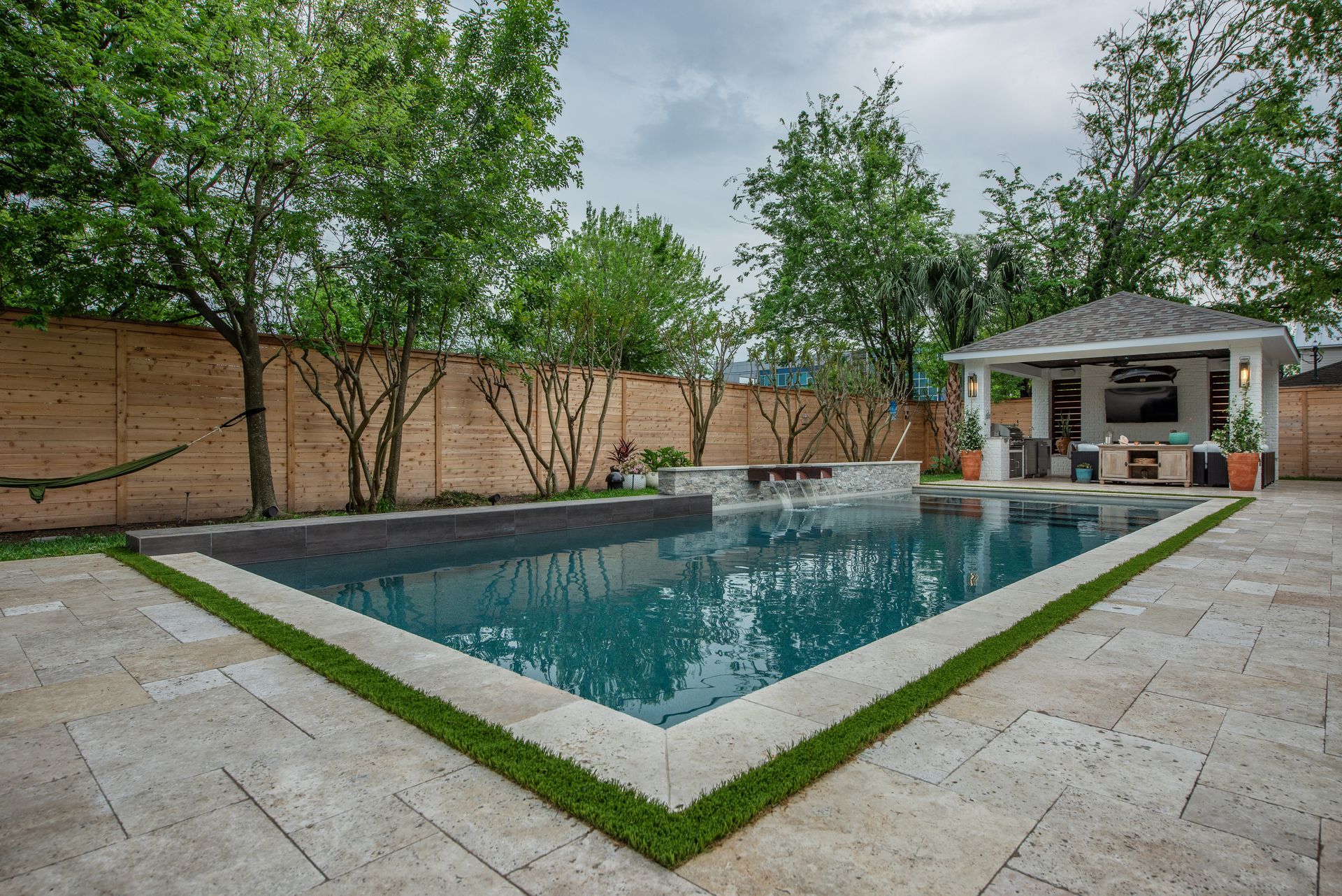 Modern backyard pool with built-in lounge chairs at sunset, surrounded by greenery and a black fence