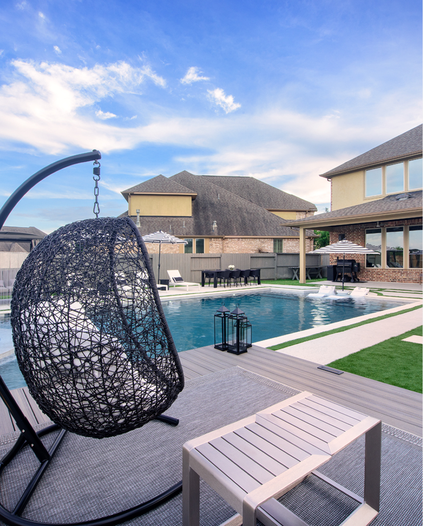 Luxury poolside patio with a hanging egg chair, lounge bench, and modern villas under a blue sky