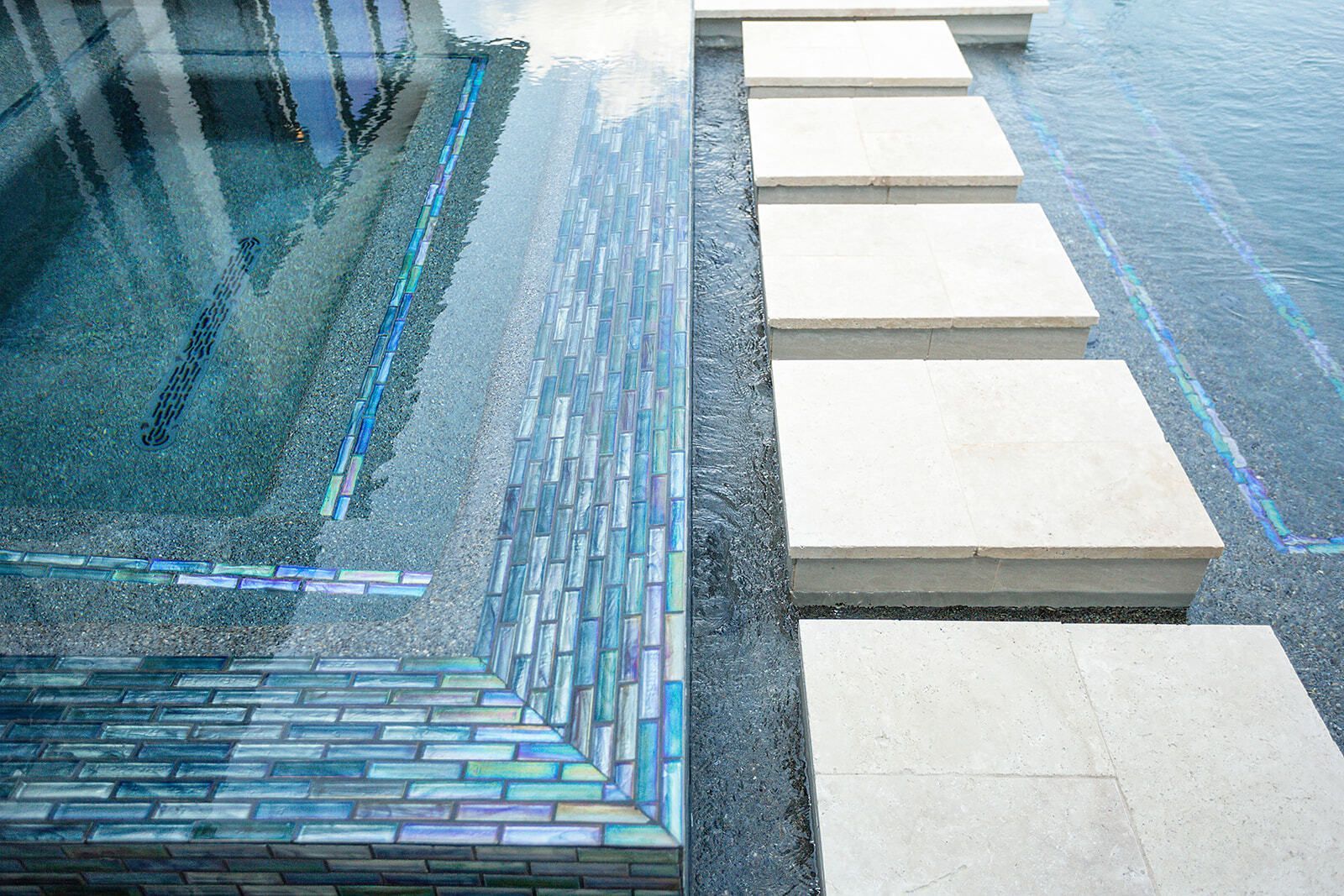 Modern outdoor pool with clear water, blue mosaic tiles, and lounge chairs beside trees under a blue sky