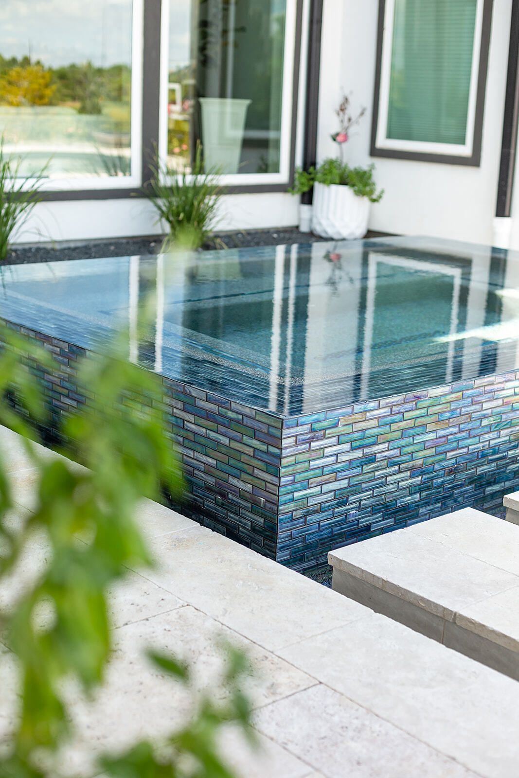 Modern outdoor pool with clear water, blue mosaic tiles, and lounge chairs beside trees under a blue sky