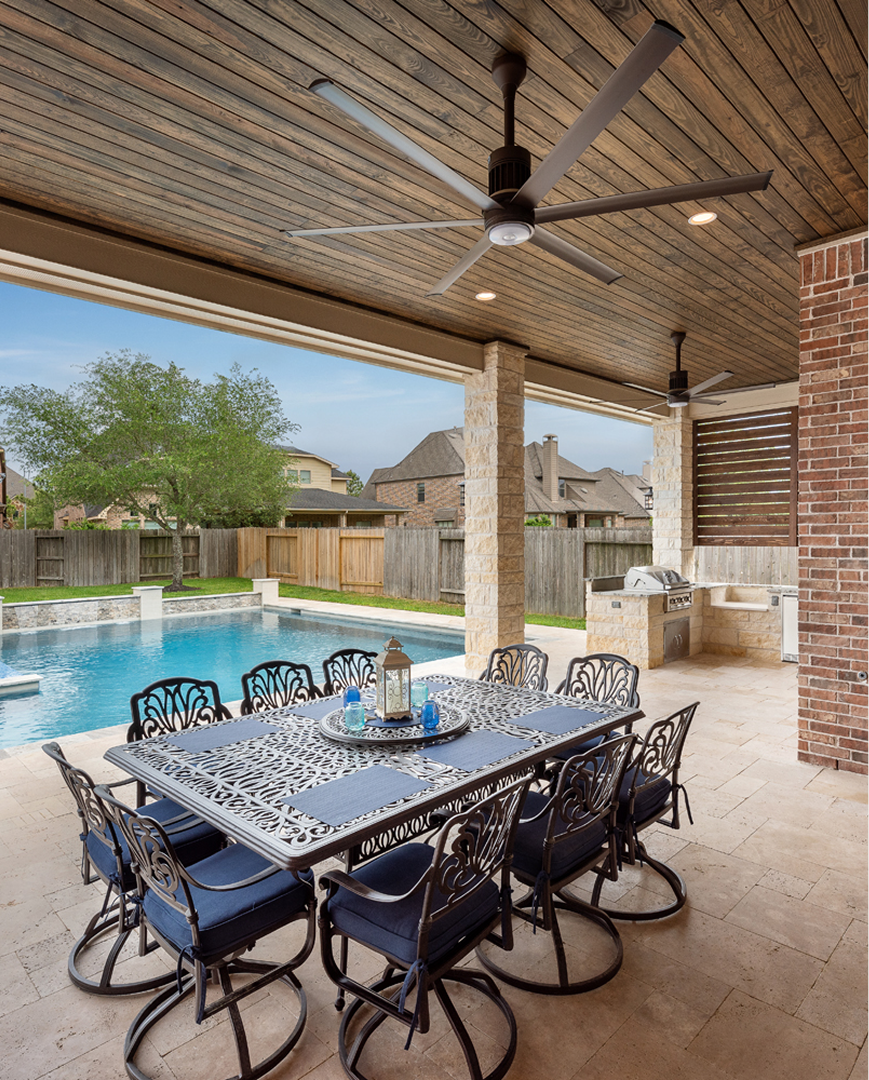 Covered patio with dining table by a backyard pool, brick columns, and ceiling fan
