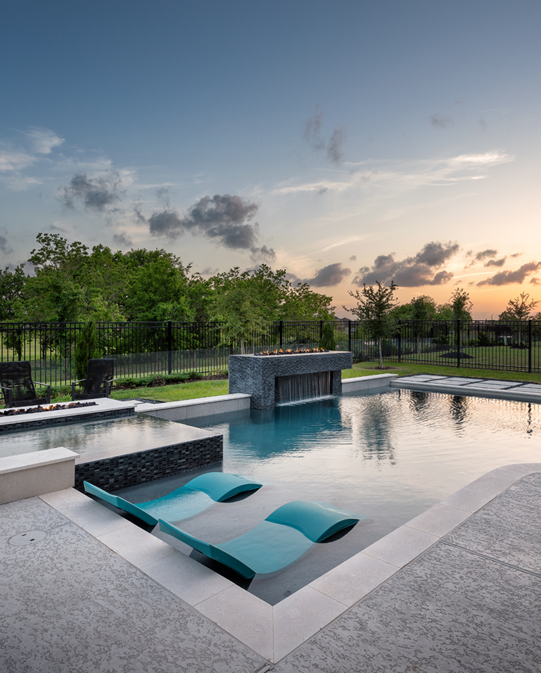 Modern backyard pool with built-in lounge chairs at sunset, surrounded by a black fence and greenery
