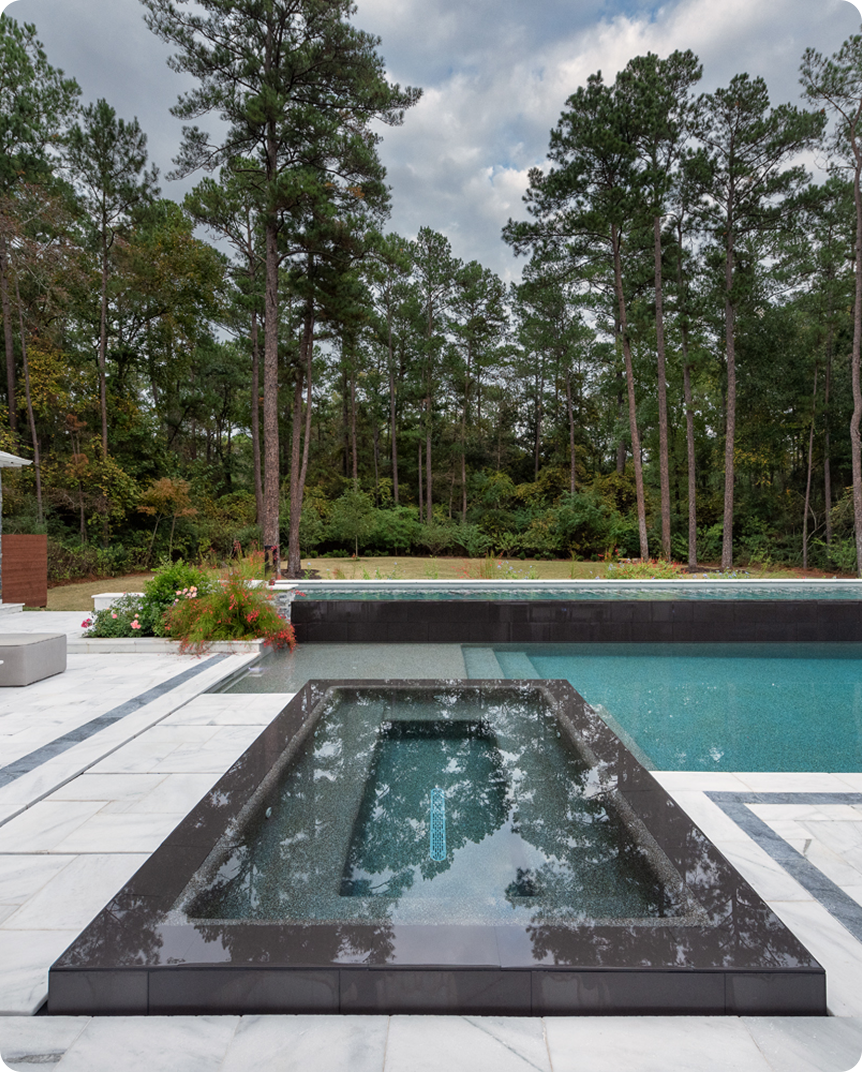 Modern backyard pool with a square hot tub in the foreground and tall pine trees beyond