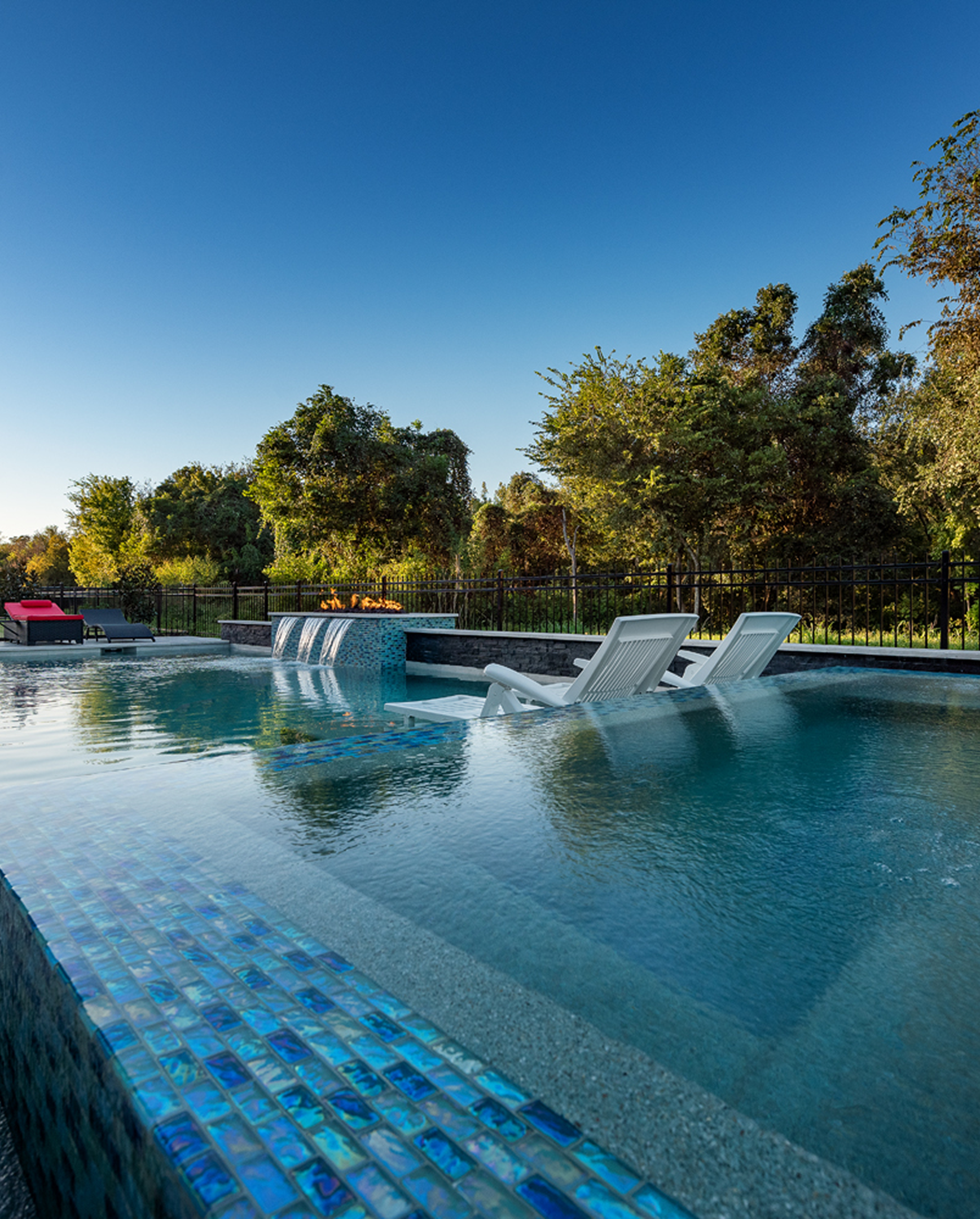 Outdoor pool with clear water, blue mosaic tiles, and lounge chairs surrounded by trees under a blue sky