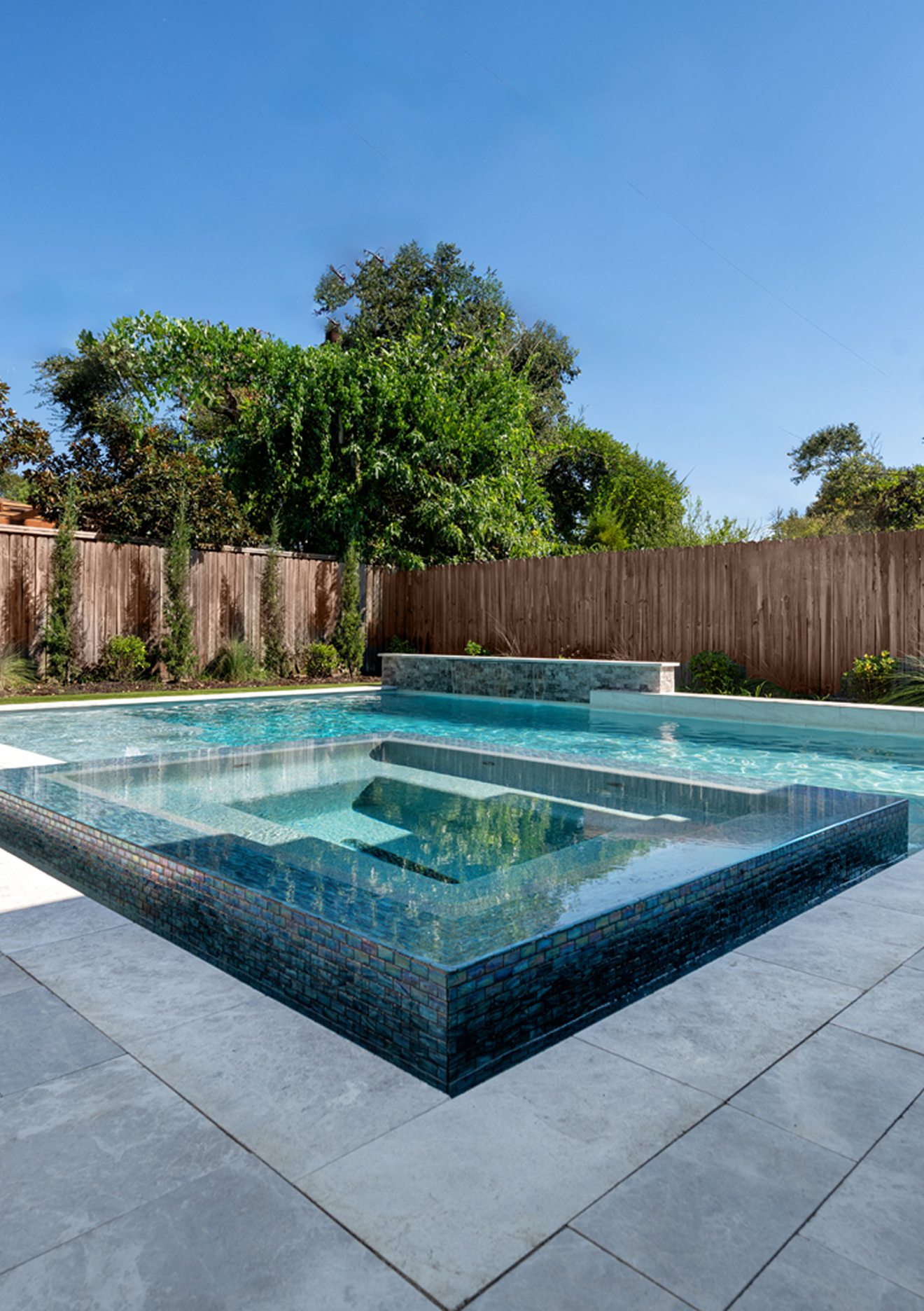 Modern backyard with a raised square pool, gray patio tiles, wooden fence, and lush greenery under a blue sky