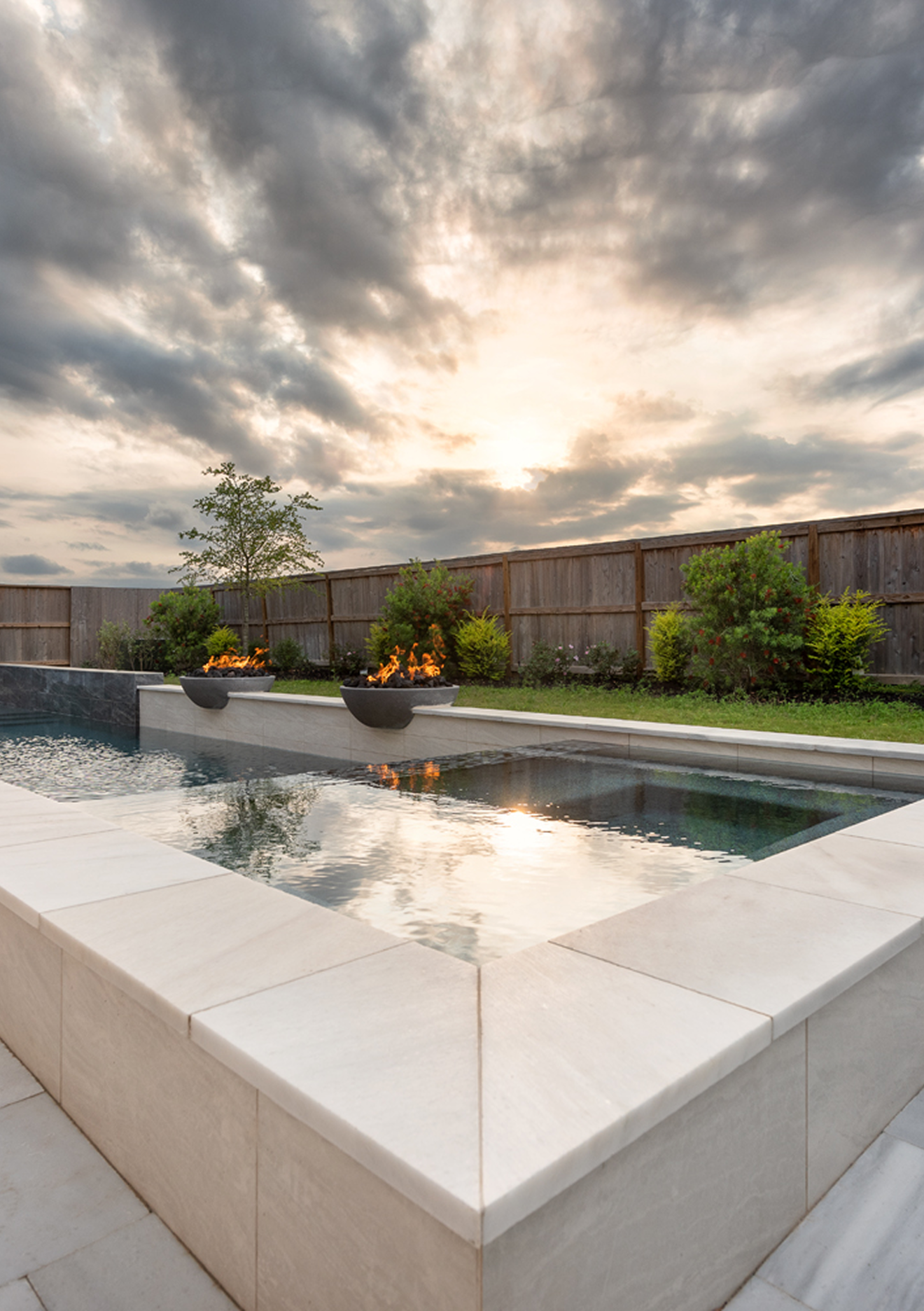 Modern backyard pool with white stone edging, reflecting cloudy sunset sky and surrounded by plants and fence