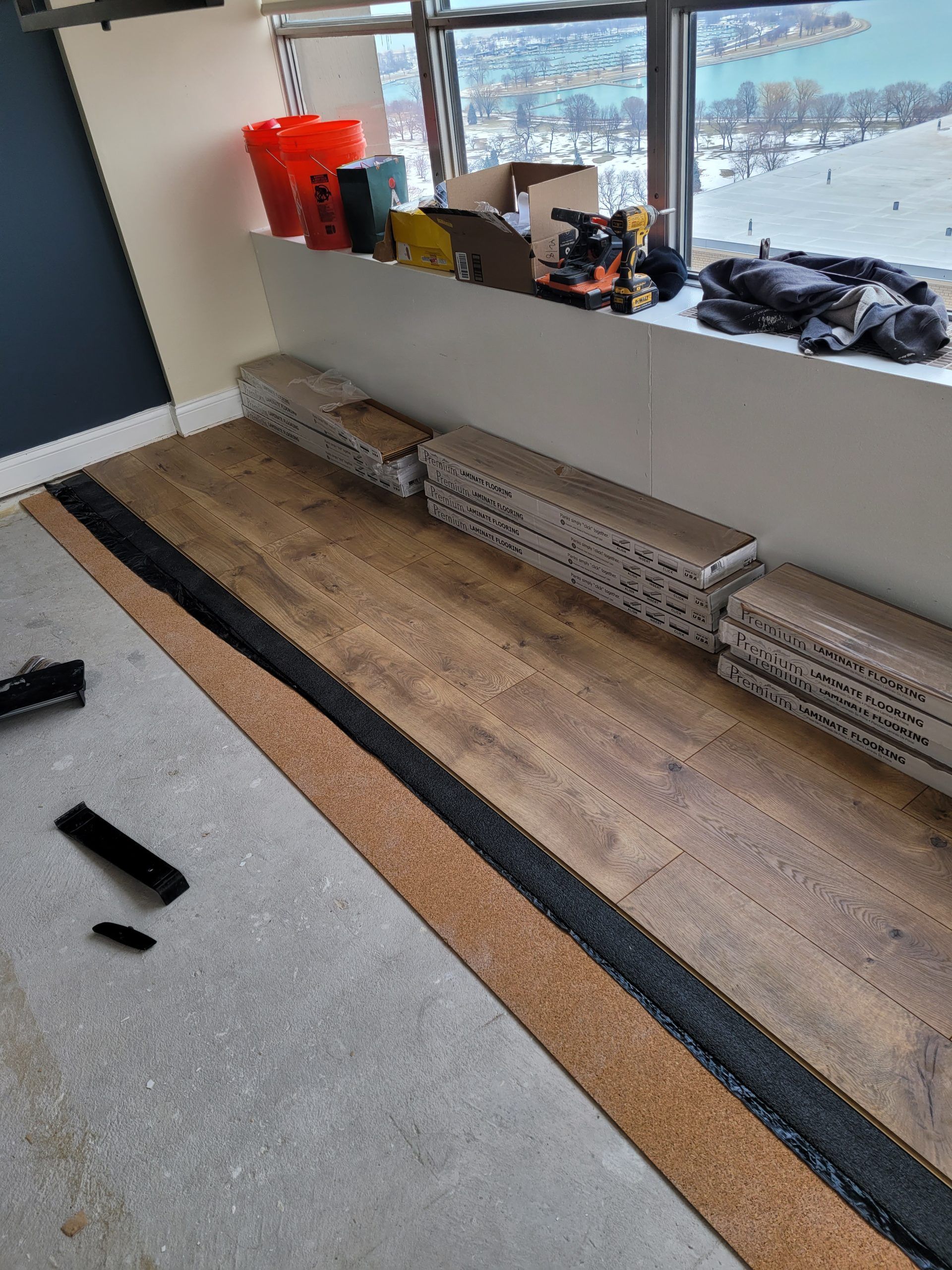 Flooring installation: stacks of wood planks, exposed cork underlayment, window with city view, tools.