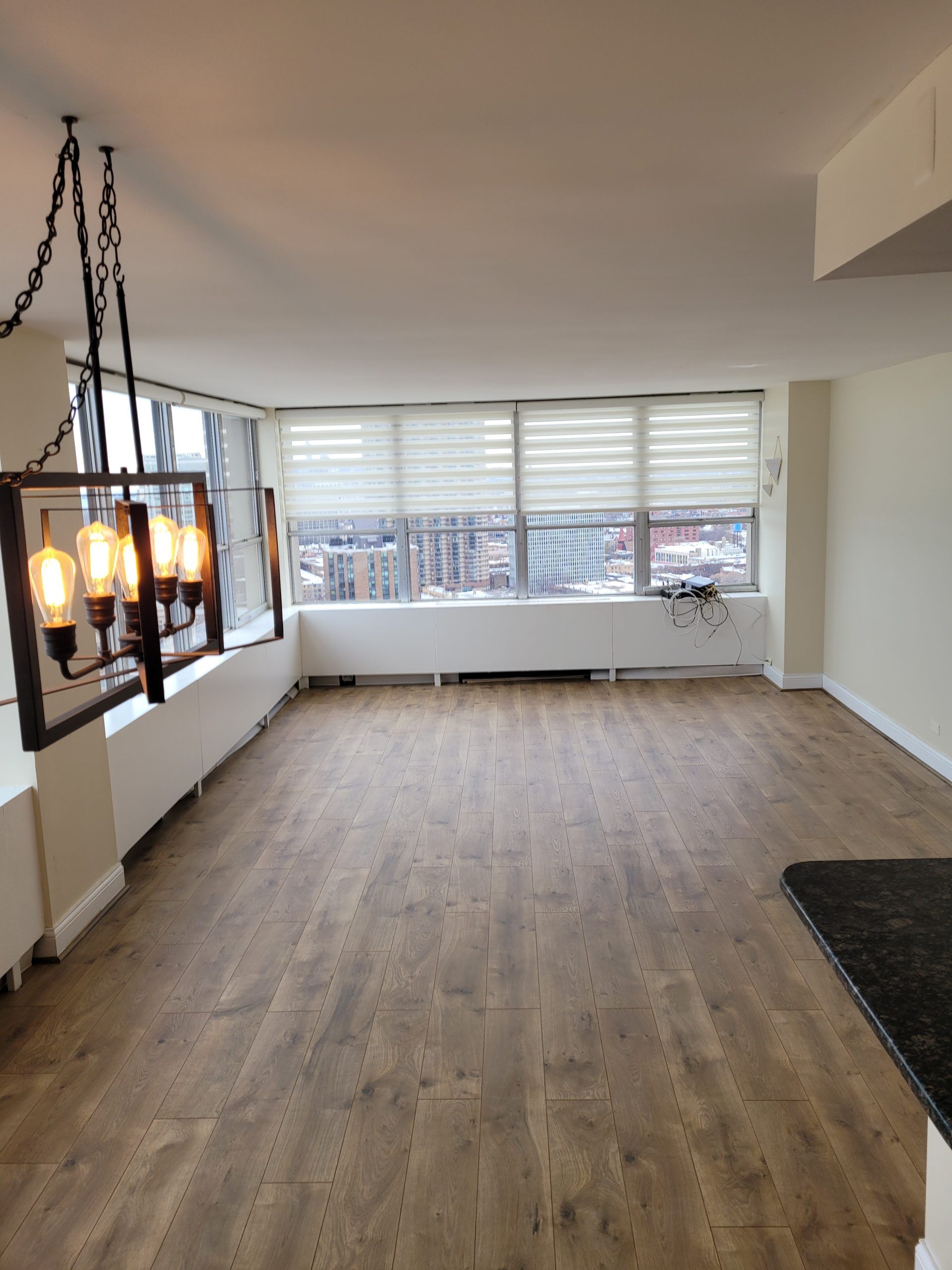 Empty living room with wood-look flooring, large windows, and a hanging light fixture.