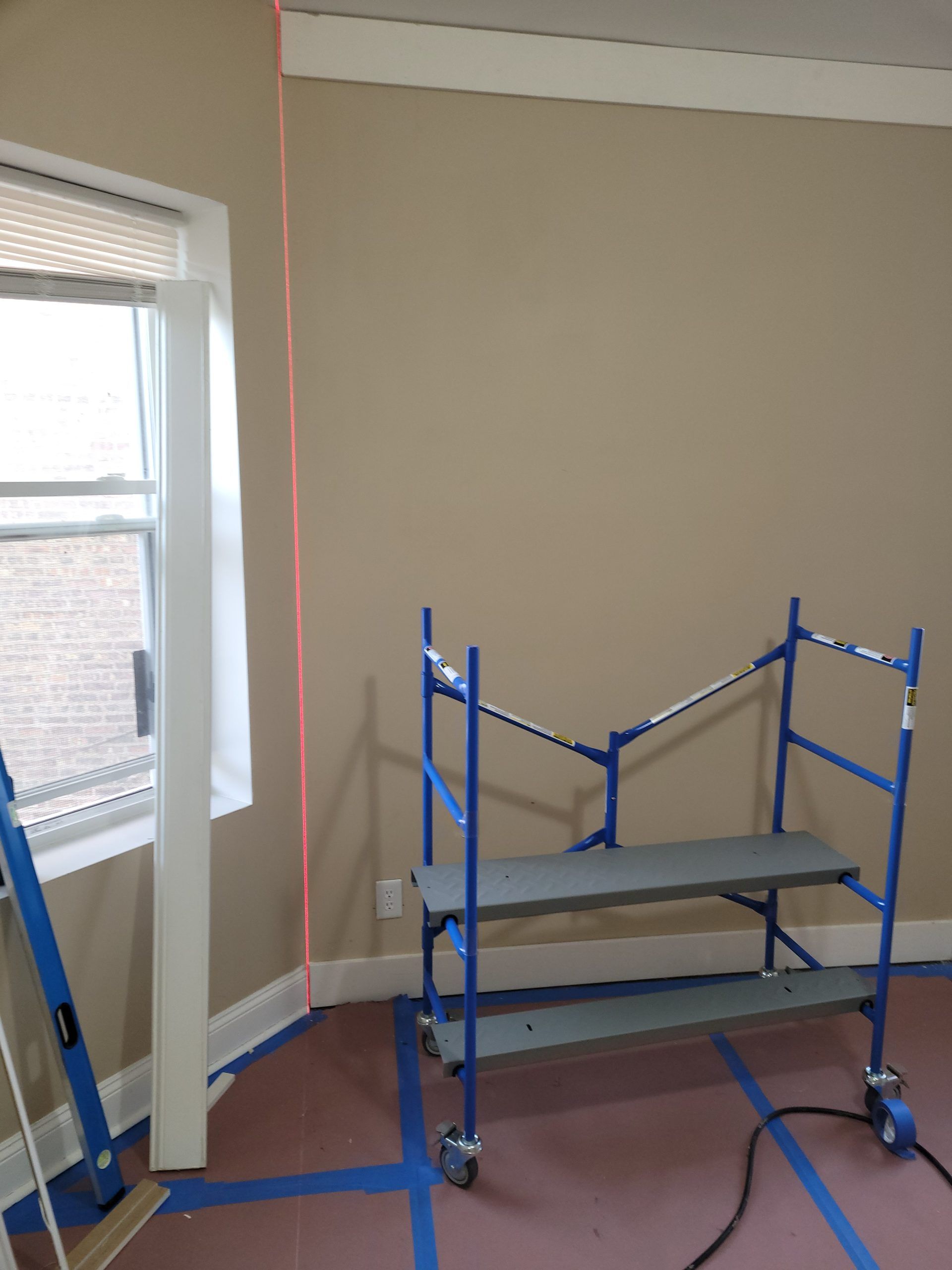 A laser level projects a red line on a tan wall. A scaffold and window are also present.
