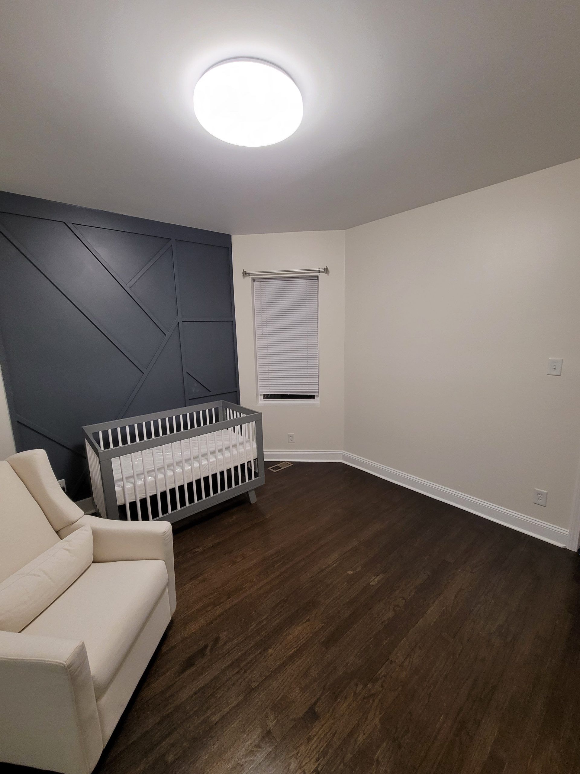 Baby nursery with crib, armchair, window, dark accent wall, and wood floors.