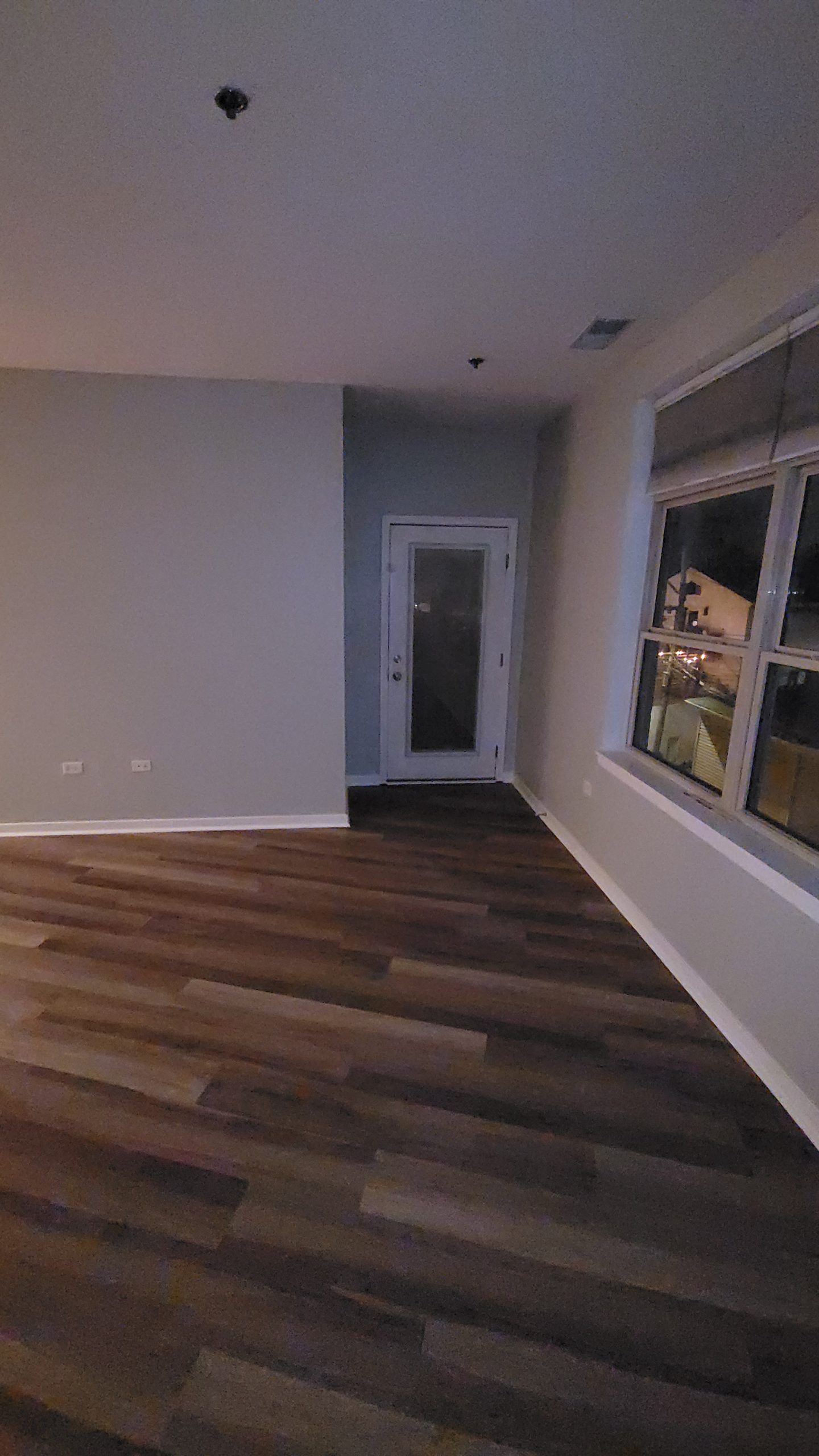 Interior view of a room with wood flooring, light-colored walls, a door, and a window with blinds.