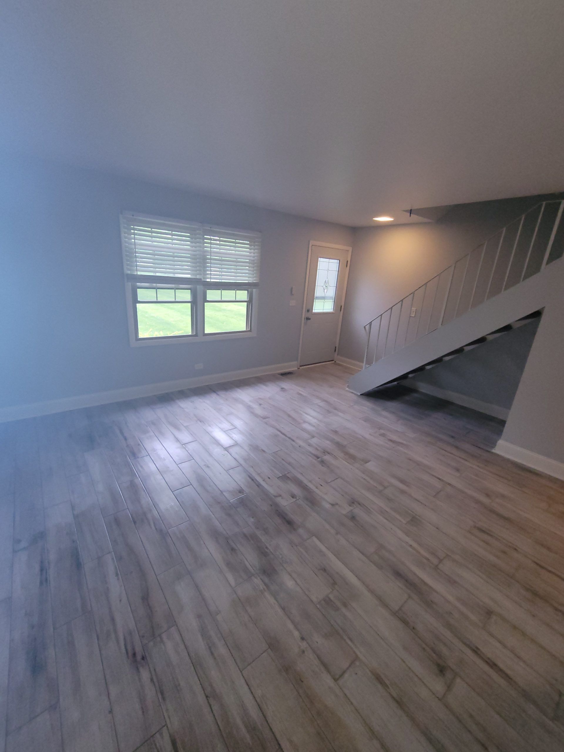 Empty room with hardwood floors, a window with blinds, and a staircase leading upwards.