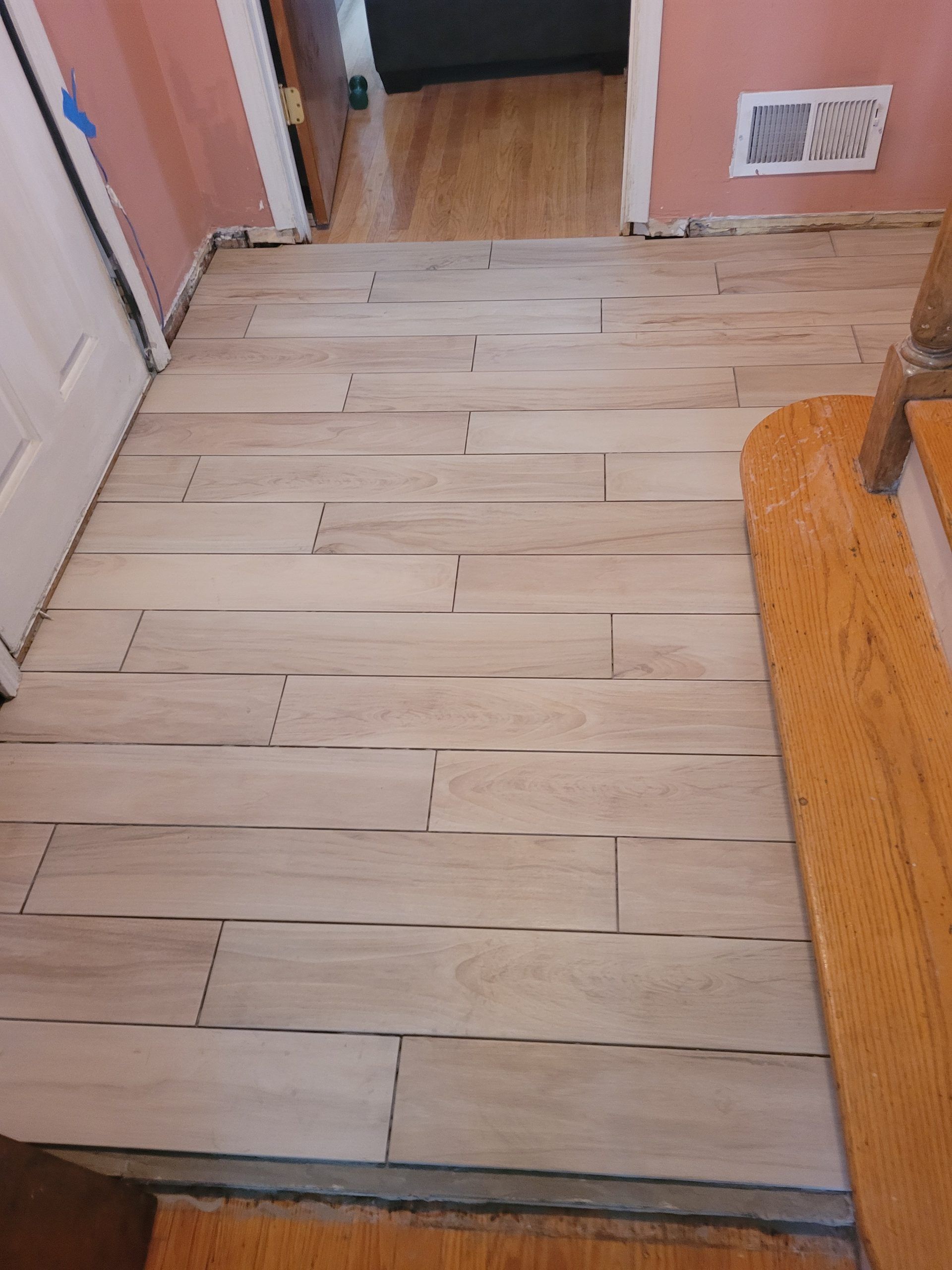 Newly installed light wood-look tile flooring in a hallway, with a door and stairs visible.
