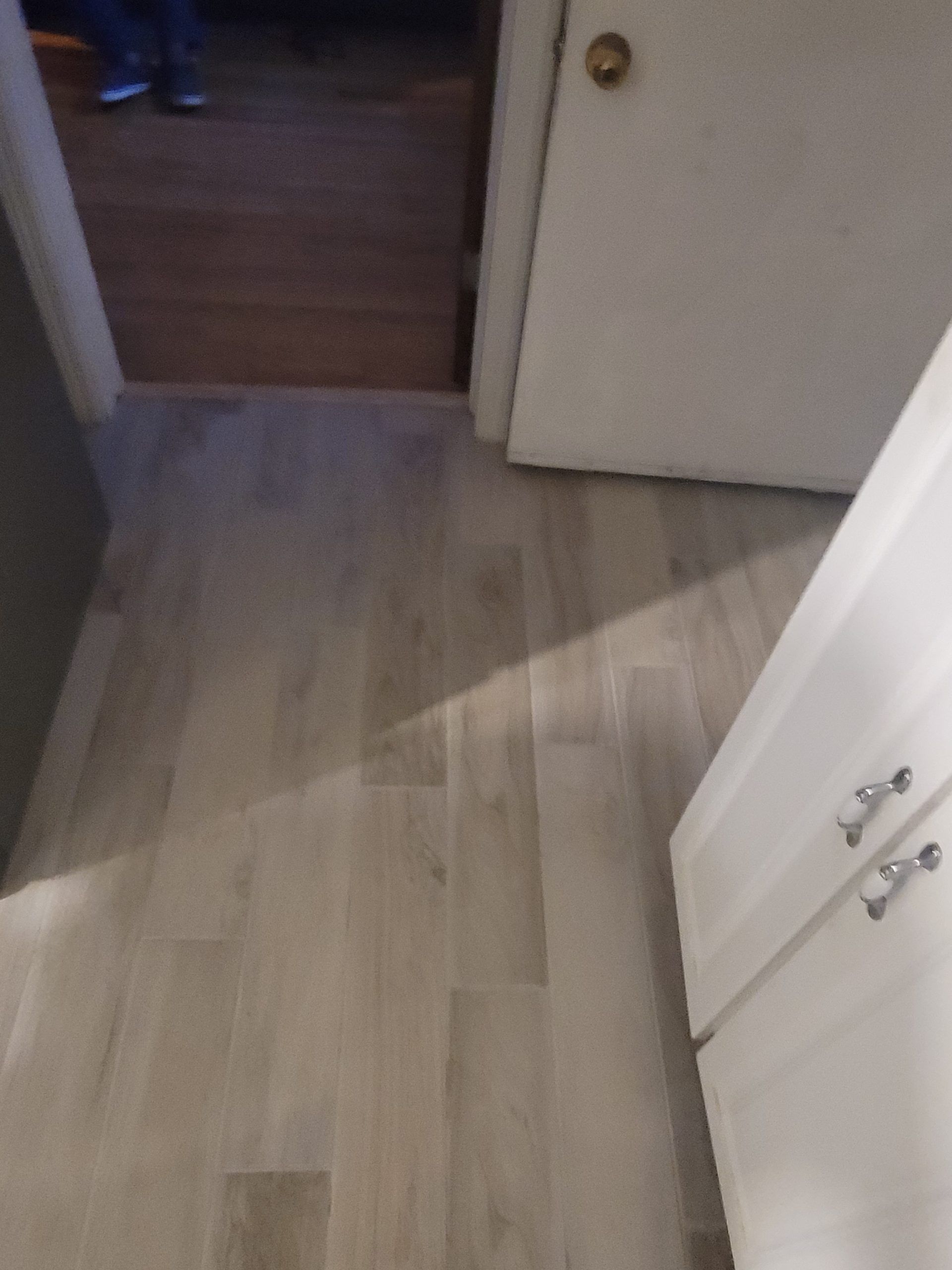 Light gray tile floor in a kitchen, with a white cabinet, door, and doorway to a wooden floor.