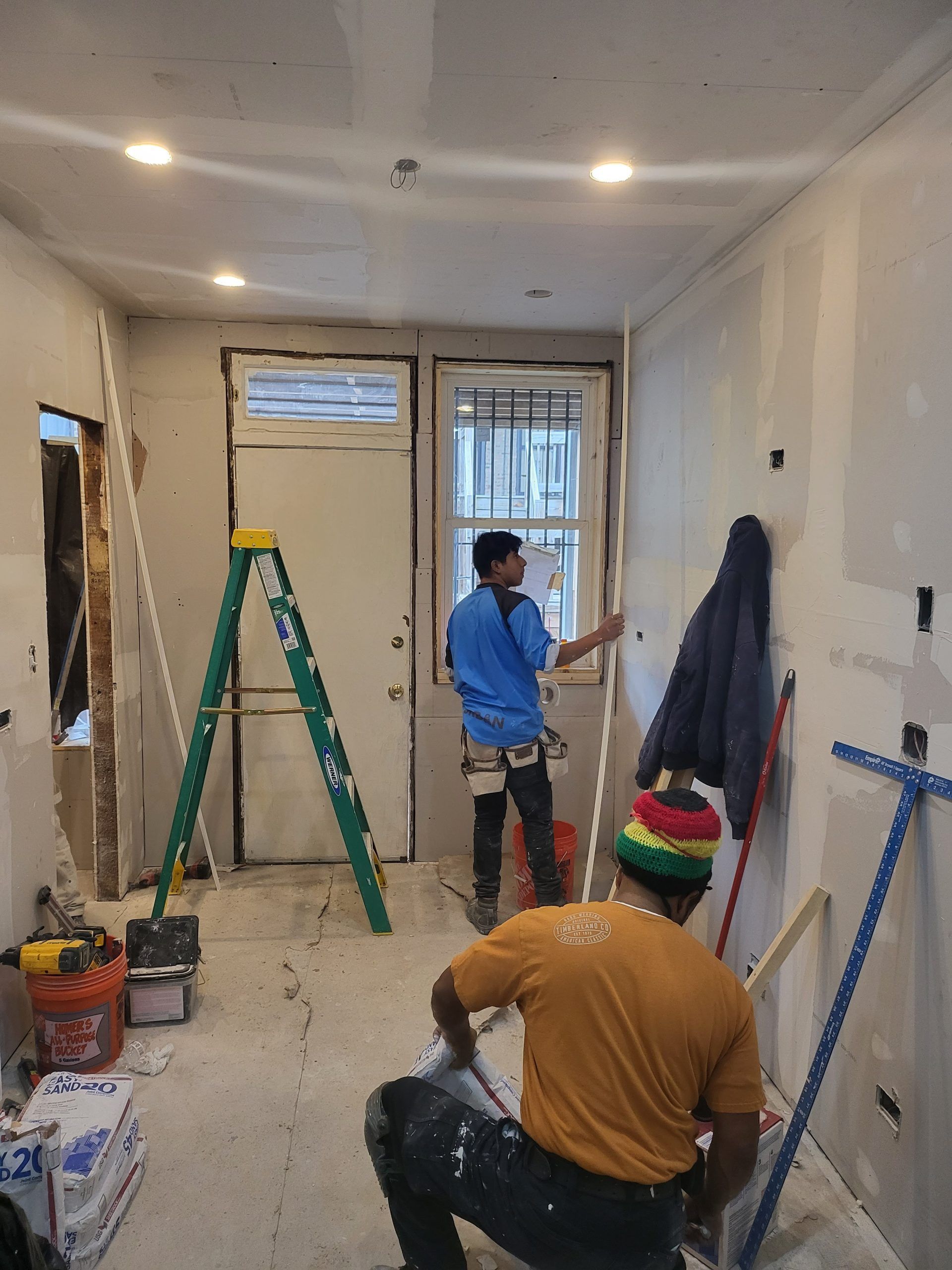 Two construction workers in a room with drywall, one applying material to the wall and the other inspecting a window.