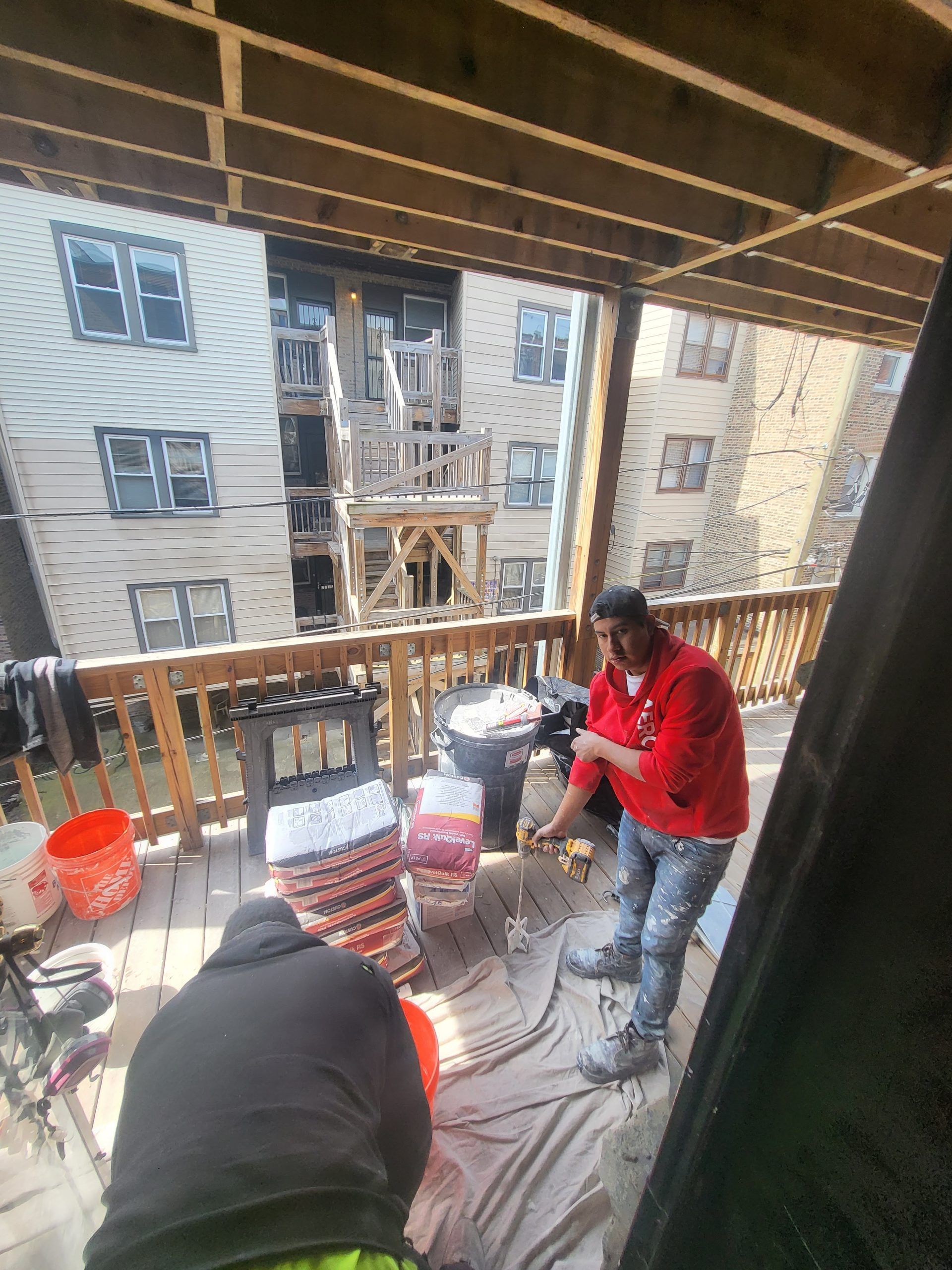 Two people on a wooden deck painting. One wears red, the other gray. Buildings in the background.