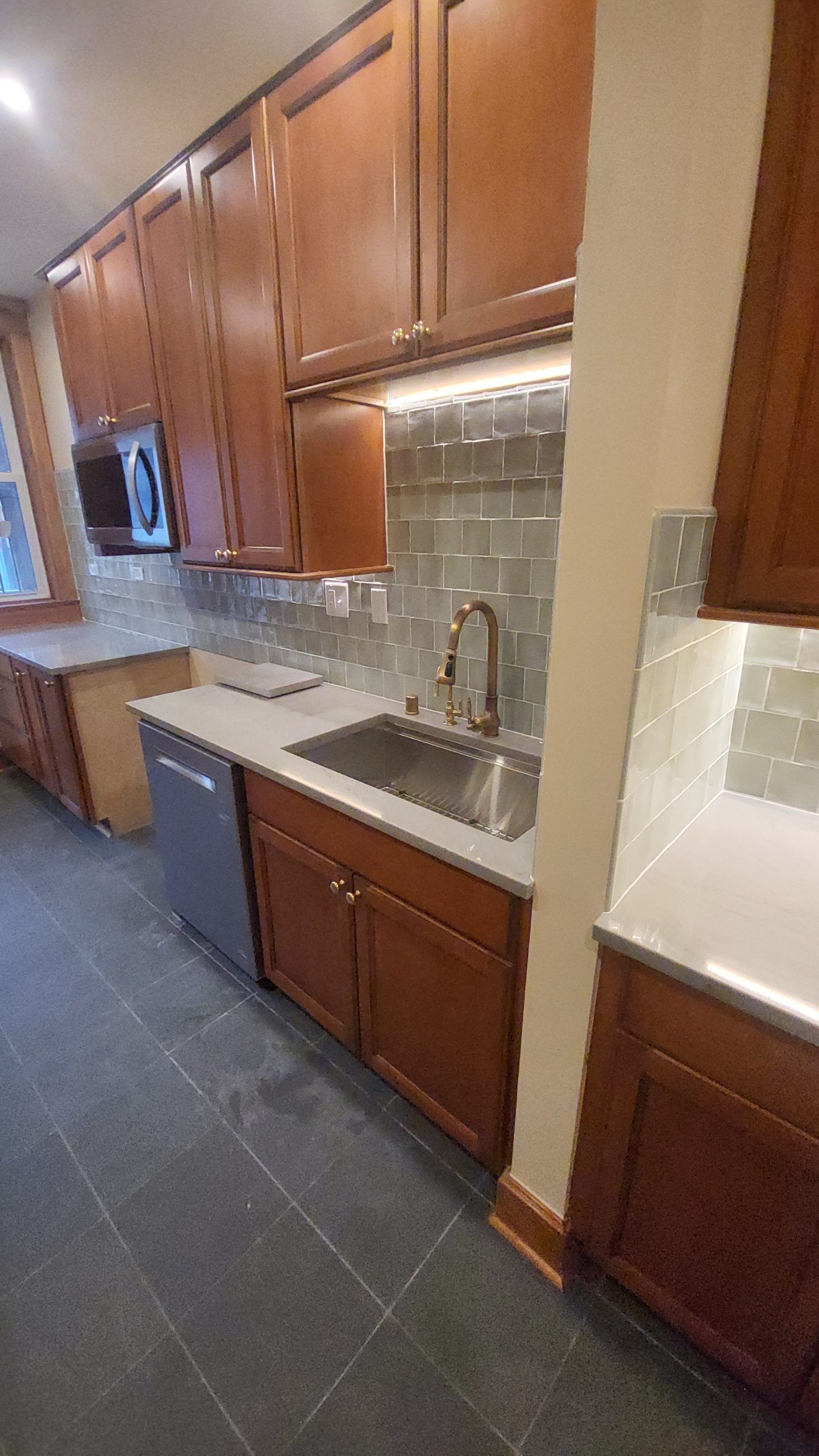 Kitchen with brown cabinets, gray backsplash, stainless steel sink, and dishwasher; gray tiled floor.