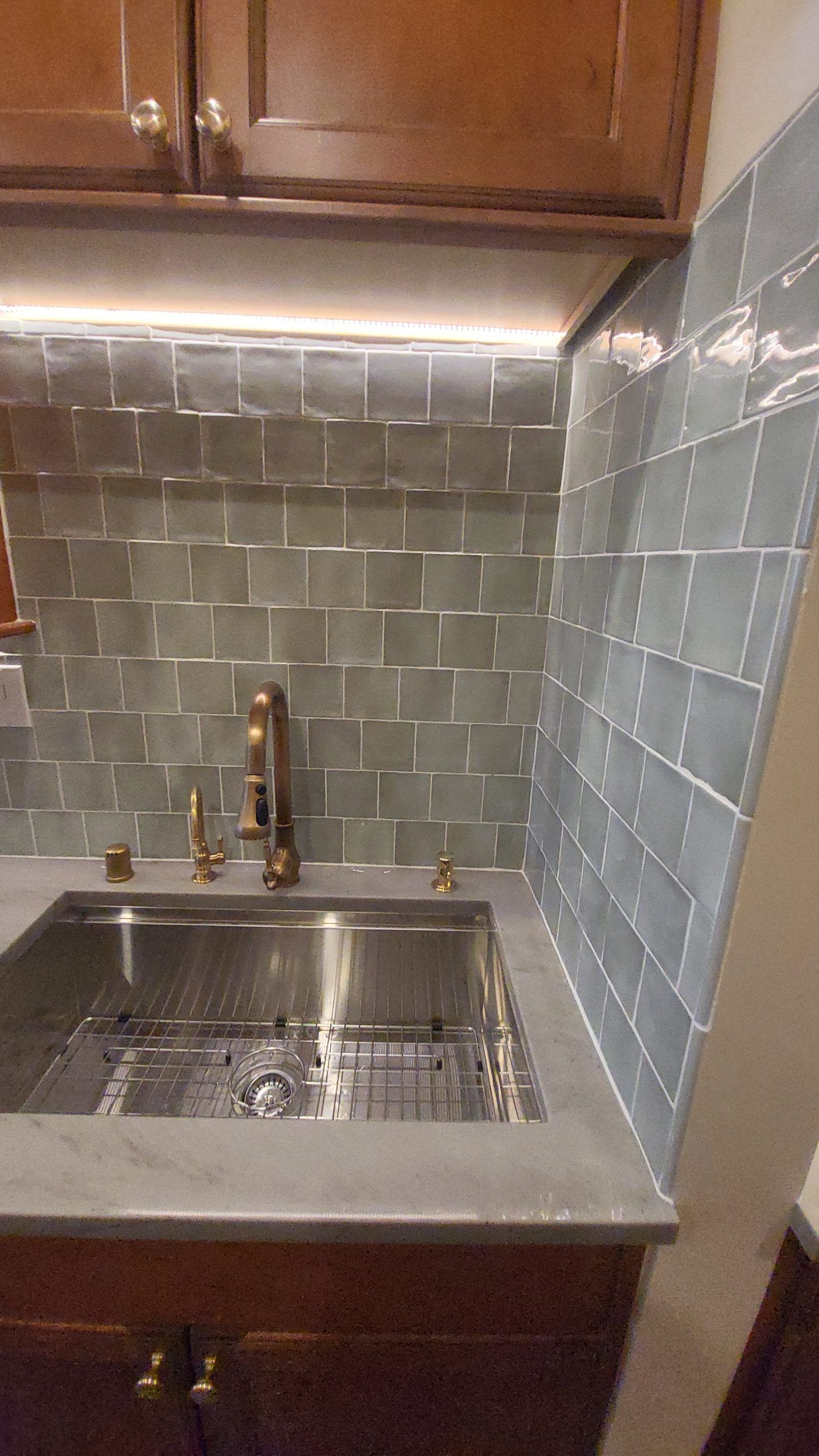 Kitchen sink with gray tile backsplash and brass faucet. Stainless steel sink set in a concrete countertop.
