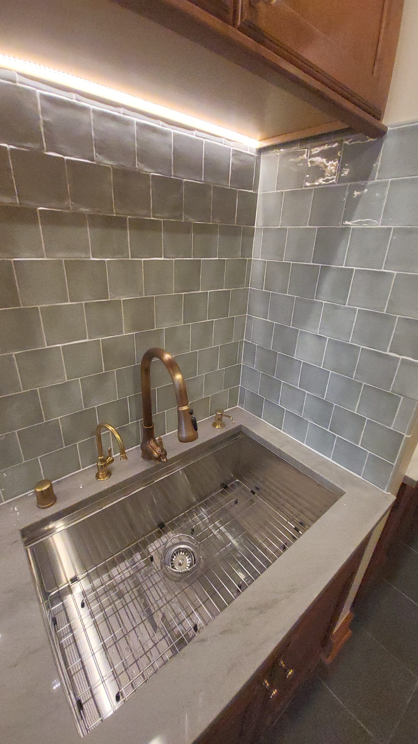 Stainless steel kitchen sink with gold faucet against grey tiled backsplash and under-cabinet lighting.